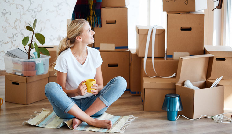 smiling woman with tea in hand sitting on floor of new apartment, pile of moving boxes on background